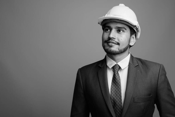 Young handsome bearded Persian businessman wearing hardhat against gray background - Foto, imagen