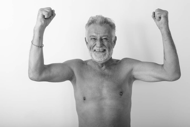 Studioaufnahme eines glücklichen älteren bärtigen Mannes, der lächelt, während er aufgeregt ohne Hemd aussieht und beide Arme vor weißem Hintergrund hebt - Foto, Bild