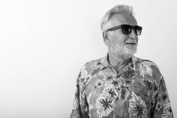 Studio shot szczęśliwego starszego brodatego turysty uśmiechającego się myśląc i nosząc okulary przeciwsłoneczne na białym tle - Zdjęcie, obraz