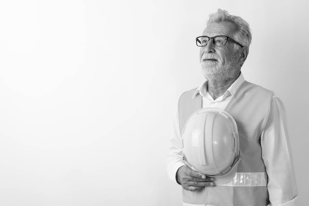 Studio shot of happy senior bearded man construction worker smiling and thinking while holding hard hat against white background - Fotoğraf, Görsel