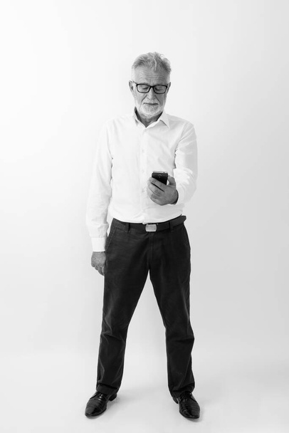 Ganzkörperaufnahme eines gutaussehenden älteren bärtigen Mannes, der sein Mobiltelefon vor weißem Hintergrund steht - Foto, Bild