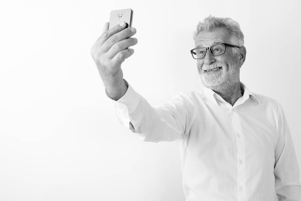 白い背景に眼鏡をかけながら携帯電話で自撮り写真を撮りながら笑顔の先輩ひげ男のスタジオ撮影 - 写真・画像