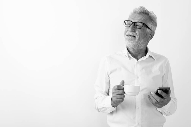Estudio de tiro de hombre barbudo feliz reflexivo sonriendo mientras sostiene la taza de café y el teléfono móvil con gafas sobre fondo blanco - Foto, Imagen