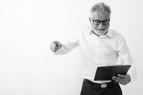 Studio shot of angry senior bearded man reading on clipboard while pointing finger with eyeglasses against white background - Photo, Image