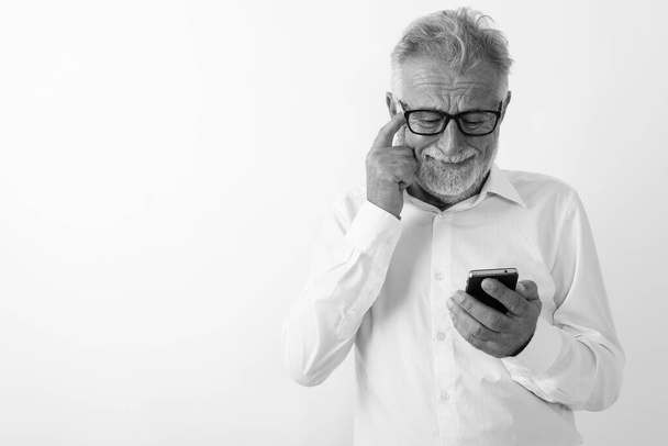 Studioaufnahme eines traurigen bärtigen älteren Mannes mit Handy, während er sich mit Brille vor weißem Hintergrund am Kopf kratzt - Foto, Bild