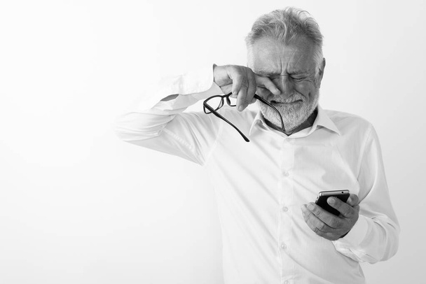Studio ujęcie smutnego starszego brodatego mężczyzny używającego telefonu komórkowego podczas płaczu i trzymającego okulary na białym tle - Zdjęcie, obraz