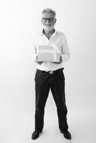 Full body shot szczęśliwego starszego brodatego mężczyzny uśmiechającego się i stojącego trzymając pudełko na białym tle - Zdjęcie, obraz