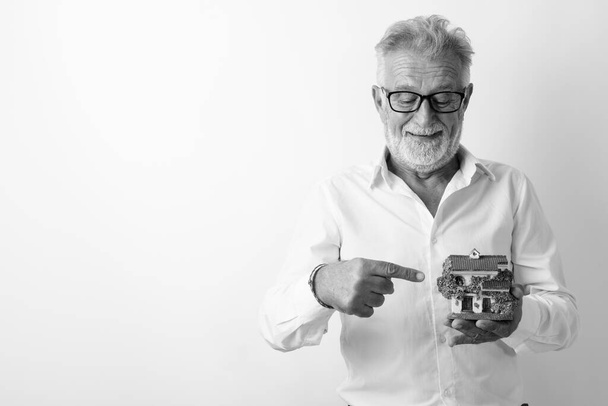 Studioaufnahme eines glücklichen älteren bärtigen Mannes, der lächelt, während er auf eine Hausfigur mit Brille vor weißem Hintergrund zeigt - Foto, Bild