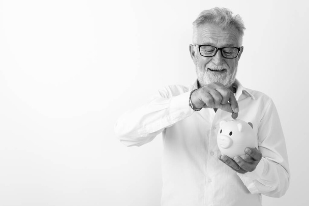 Studio shot of happy senior bearded man smiling while looking and putting coin in piggy bank with eyeglasses against white background - Фото, изображение