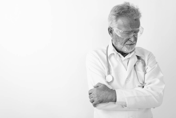Studio shot of sad senior bearded man doctor thinking while wearing protective glasses with arms crossed against white background - Foto, afbeelding