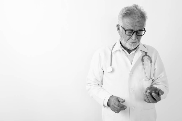 Studio shot of handsome senior bearded man doctor looking at tomato while wearing eyeglasses against white background - Foto, afbeelding