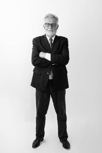 Foto de cuerpo completo de feliz hombre de negocios barbudo mayor sonriendo y de pie mientras lleva gafas con los brazos cruzados sobre fondo blanco - Foto, Imagen