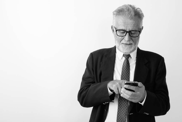 Studio shot of handsome senior bearded businessman using mobile phone while wearing eyeglasses against white background - Photo, image