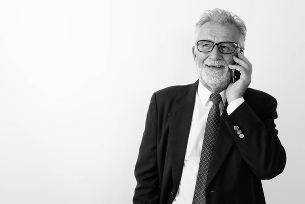 Studio shot szczęśliwego starszego brodatego biznesmena uśmiechającego się myśląc i rozmawiając przez telefon komórkowy na białym tle - Zdjęcie, obraz
