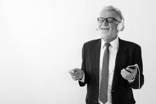 Студійний знімок вдумливого щасливого старшого бородатого бізнесмена, посміхаючись, тримаючи мобільний телефон і слухаючи музику на білому тлі
 - Фото, зображення