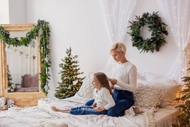 Mother braids her daughter's braids in bed. Morning Christmas calmness relax cozy. New Year's decor in the bedroom. Xmas mood. Weekend leisure togetherness. Motherhood childhood. white sweater - Valokuva, kuva