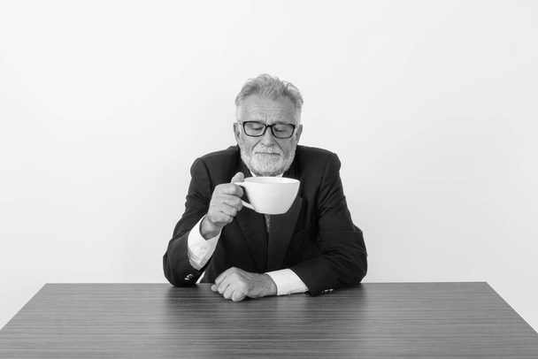 Студійний знімок красивого старшого бородатого бізнесмена тримає і дивиться на чашку кави, сидячи на дерев'яному столі на білому тлі
 - Фото, зображення