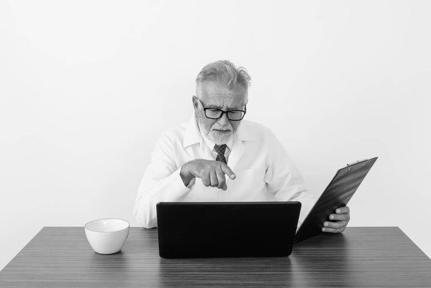 Studio strzał przystojny starszy brodaty mężczyzna lekarz trzymając schowek wskazując na laptopa na drewnianym stole na białym tle - Zdjęcie, obraz