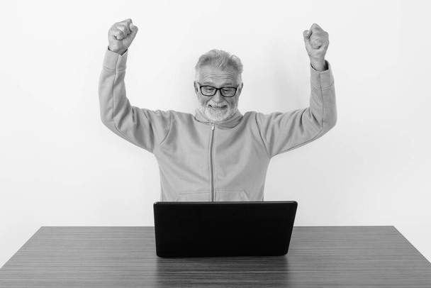 Heyecanlı görünürken gülümseyen ve tahta masada beyaz arka plana karşı dizüstü bilgisayar kullanan yaşlı ve mutlu bir adamın stüdyo fotoğrafı. - Fotoğraf, Görsel