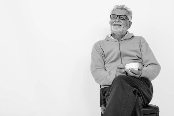 Studio záběr zamyšlený šťastný senior vousatý muž s úsměvem při držení šálek kávy a posezení na židli připraven pro tělocvičnu proti bílému pozadí - Fotografie, Obrázek