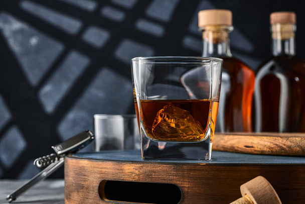 Square glass of whiskey with ice on a wooden tray, on a blue background. Two glasses of brandy and ice on a wooden tray, on a blue background. - Zdjęcie, obraz