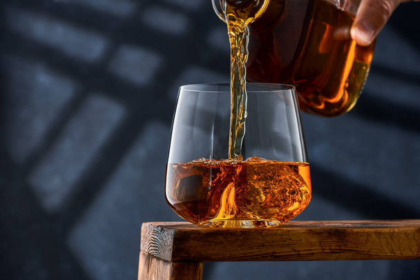 Crystal glass of whiskey with ice on a wooden counter, in a glass of whiskey poured from a bottle. A glass of brandy with ice on a wooden counter, brandy is poured into the glass from a bottle. - Photo, image
