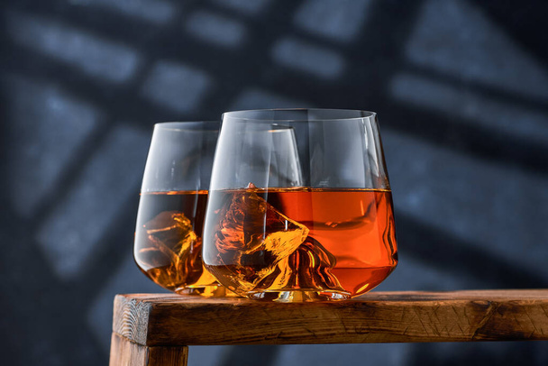 Two crystal glasses of whiskey with ice on a wooden counter on a blue background. Two crystal glasses with a double portion of brandy on a wooden counter on a blue background. - Photo, image