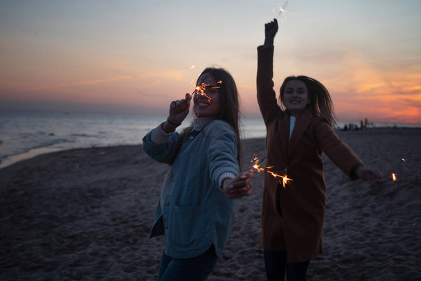 Two cheerful friends on the beach in the evening light sparklers. - Φωτογραφία, εικόνα