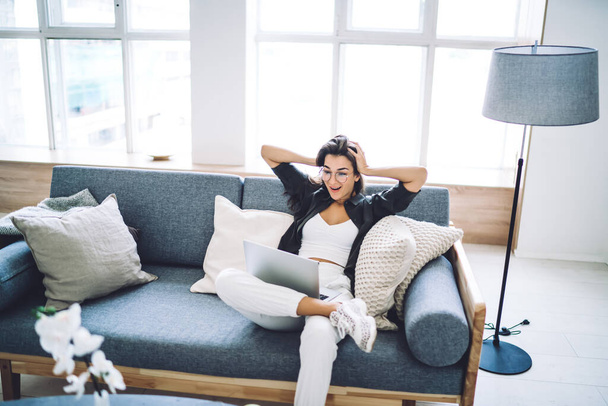 From above of young cheerful woman in casual clothes and eyeglasses sitting on comfortable sofa and browsing laptop while spending time time on quarantine - 写真・画像