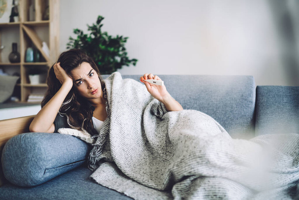 Exhausted caucasian hipster girl resting on sofa feeling ill and fever getting respiratory virus during influenza, pandemic,sad depressive woman suffer from viral infection checking temperature via termometr - Fotoğraf, Görsel