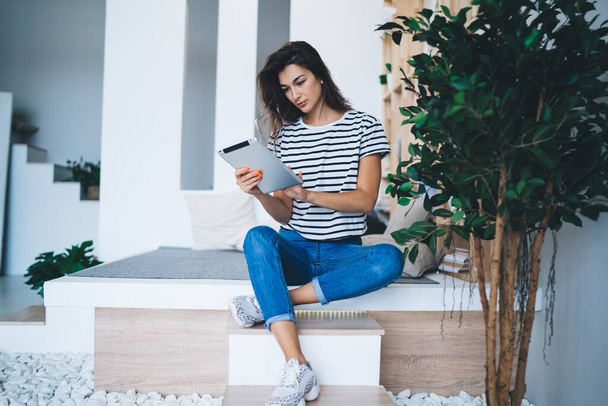 Concentrated young female freelancer in casual outfit sitting on cozy podium in contemporary apartment and browsing tablet while working remotely - Фото, зображення