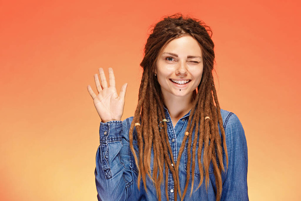 Funny and joyful winking young woman with dreadlocks and greeting gesture - 写真・画像