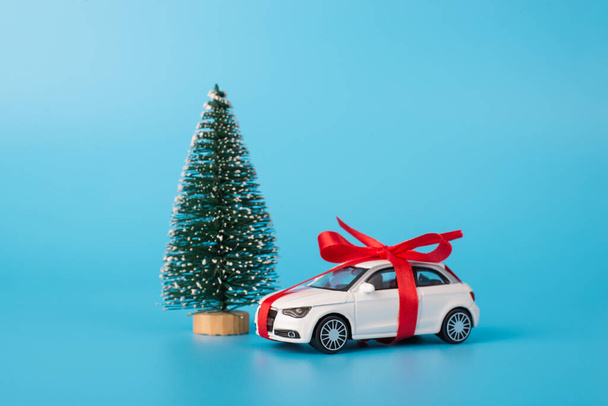 Gewenste aanwezig op kerstconcept. Close-up foto van klein speelgoed luxe stijlvolle auto verpakt met rood lint staan in de buurt van sparren groene boom geïsoleerd over lichtkleur blauwe achtergrond - Foto, afbeelding