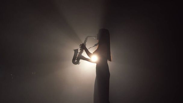 Молода красива дівчина в темній сукні грає на золотому блискучому саксофоні на сцені. Темна студія з димом і освітленням сцени. Руки і саксофон крупним планом. Вид збоку. Низьке відео руху
 - Фото, зображення