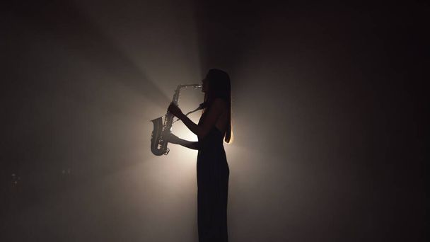 Молодая красивая девушка в темном платье играет на золотом блестящем саксофоне на сцене. Темная студия с дымом и подсветкой сцены. Руки и саксофон закрываются. Боковой просмотр видео замедленного действия - Фото, изображение