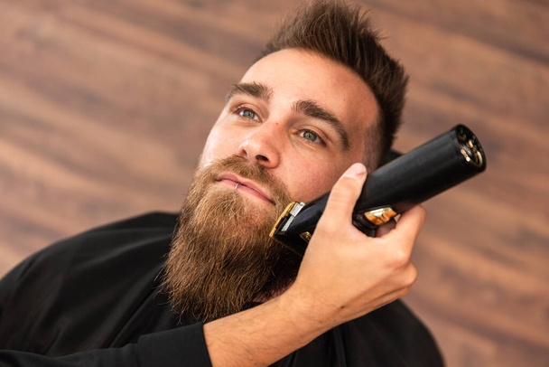 View from above of a customer in a barber shop, his beard is being trimmed and cut with an electric razor - Photo, Image
