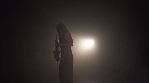 Das junge hübsche Mädchen im dunklen Kleid spielt auf einem goldglänzenden Saxophon auf der Bühne. Dunkles Studio mit Rauch und Bühnenbeleuchtung. Hände und Saxofon hautnah. Seitenansicht.Zeitlupenvideo - Foto, Bild