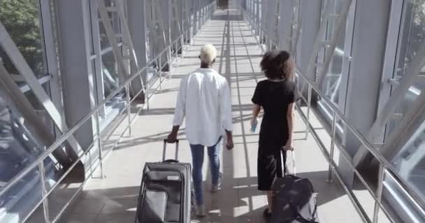 Вид сзади пары молодых африканцев, девушки-подруги муж и жена, гуляющие в терминале аэропорта с чемоданами с багажом, вместе идут на посадку, путешествуют отпуск - Кадры, видео