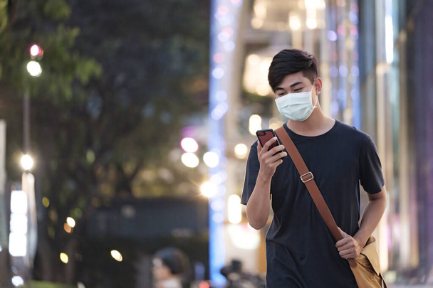 Młody człowiek w masce ochronnej przy użyciu telefonu komórkowego w centrum miasta zapobiega rozprzestrzenianiu się wirusów w mieście. Nowa koncepcja normalna. - Zdjęcie, obraz