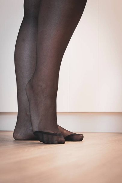Ein elegantes Porträt der Füße eines Mädchens in schwarzer Strumpfhose mit dem Nylon an den Zehen, das auf einem Holzboden steht und posiert. - Foto, Bild