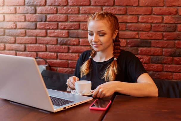Ginger girl uses laptop in cafe, sitting on sofa, wearing black casual t shirt, woman looks at laptop screen, freelancer has break from working, enjoying hot beverage, female poses against brick wall - Photo, image