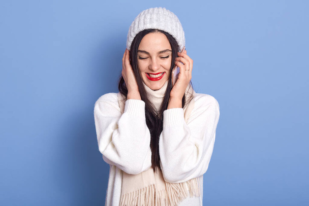 Happy pretty brunette girl talking on phone and laughing, holding smartphone near ear, female dresses white sweater, scarf, cap, standing isolated over blue background, girl wears red lipstick. - Valokuva, kuva