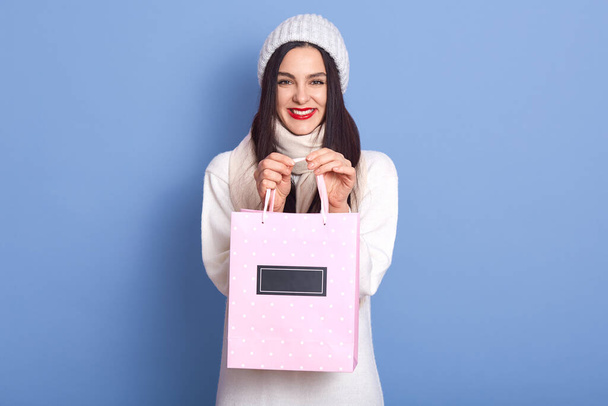 Beautiful lady indicate showing pack, has perfect look, buys present for birthday, wear white sweater, scarf and cap, posing isolated over blue background, being happy after shopping. - Foto, Imagem