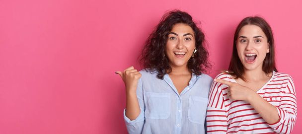 Portrait of astonished women pointing fingers aside at copy space isolated over pink background, surprising females with surprised facial expressions, keeping mouths opened. - Foto, imagen