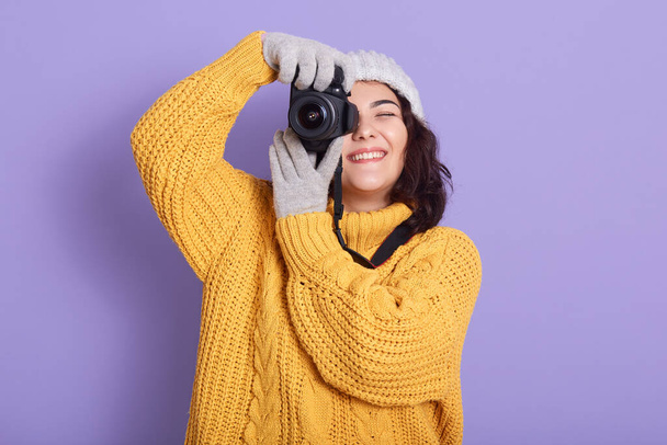 Woman takes photos, smiling female holding photographic camera, keeps one eye closed, wearing warm clothing, expressing positive emotions, standing isolated over lilac wall. - Foto, Imagem