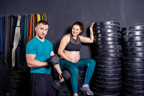 Спортсмены беременная женщина и ее муж позируют в спортзале возле спортивного инвентаря. Активная спортивная беременность. Тренировка в спортзале вместе во время беременности. Беременная женщина с мужем, занимающаяся спортом - Фото, изображение