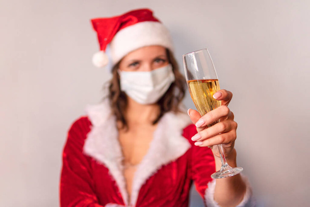 Retrato de una hermosa mujer sonriente con sombrero de navidad y vestido sosteniendo una copa de champán mientras está de pie sobre un fondo gris. La mujer con la máscara protectora del virus, Navidad en cuarentena - Foto, Imagen