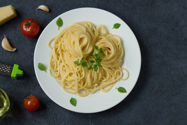 Spaghettis italiens traditionnels sur une assiette ronde blanche sur un fond sombre. Ingrédients pour pâtes - tomates cerises, parmesan, ail, huile d'olive, basilic. Vue du dessus. - Photo, image