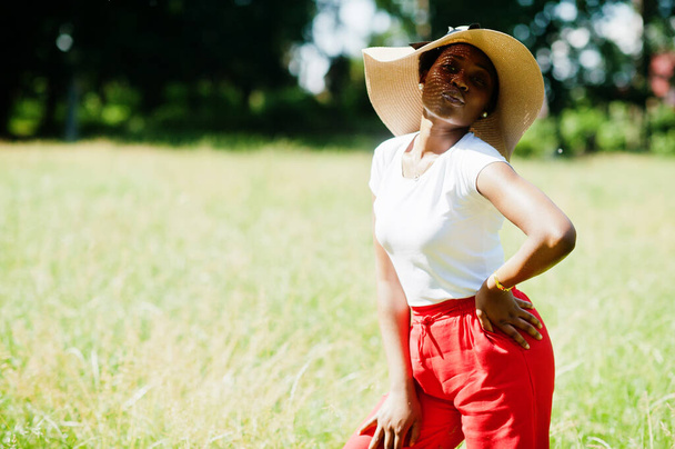 Portrait of gorgeous african american woman 20s wear in summer hat, red pants and white t-shirt, posing at green grass in park. - Φωτογραφία, εικόνα