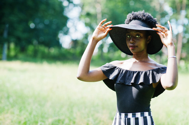 Portrait of gorgeous african american woman 20s in wear in black and white stripe pants and summer hat posing at green grass in park. - Foto, Bild